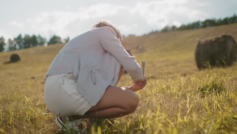 female tourist squatting while capturing a photograph of someone in a vast farmland, sunlight highlights golden grass, rolling hills, and rustic countryside scenery