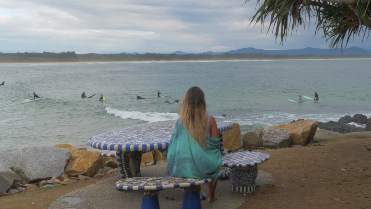 피크닉 테이블에서 휴식을 취하는 여성 뒤에서 바다에서 파도를 즐기는 서퍼 - 호주 뉴 사우스 웨일즈 주 스콧 헤드에서 서핑