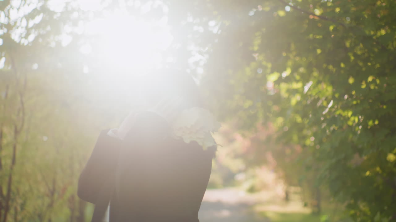 Medium shot of carefree walker in autumn jacket wearing headset, moving rhythmically and spinning round with joy while strolling along scenic forest pathway under warm sunlight