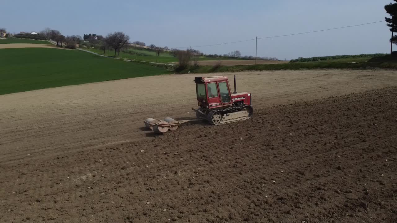Drone follow red tractor rolling the soil creating better seedbed for germinating seeds, aerial agricultural farming natural soil land