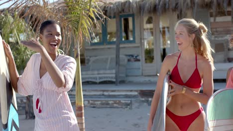 Happy-diverse-female-friends-walking-with-surfboards-and-talking-over-beach-house