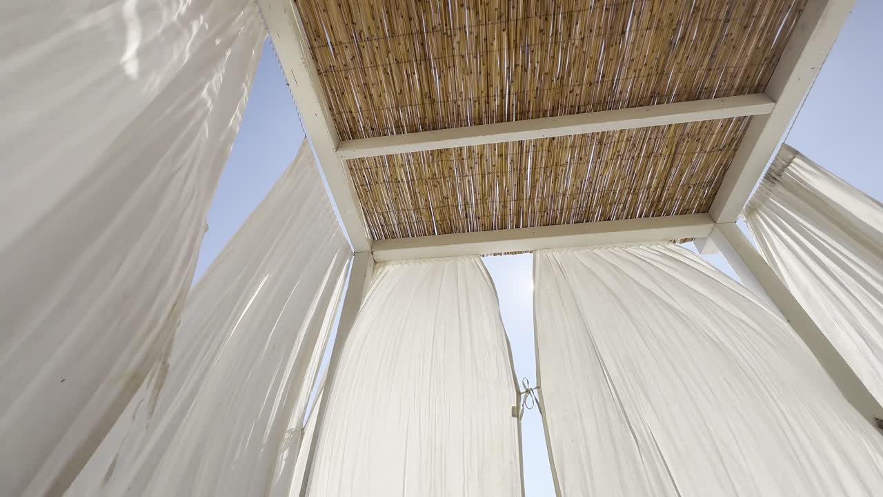 White curtains gently sway in the wind beneath a bamboo-roofed beach cabana. Bright blue sky and natural textures create a serene, tropical vacation ambiance