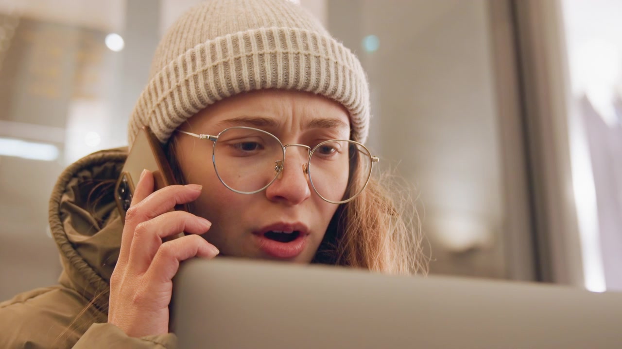 Young woman with laptop making urgent call, beanie and glasses, winter coat, tense expression while troubleshooting account issue in cozy indoor setting, mouth open speaking, eyes focused on screen