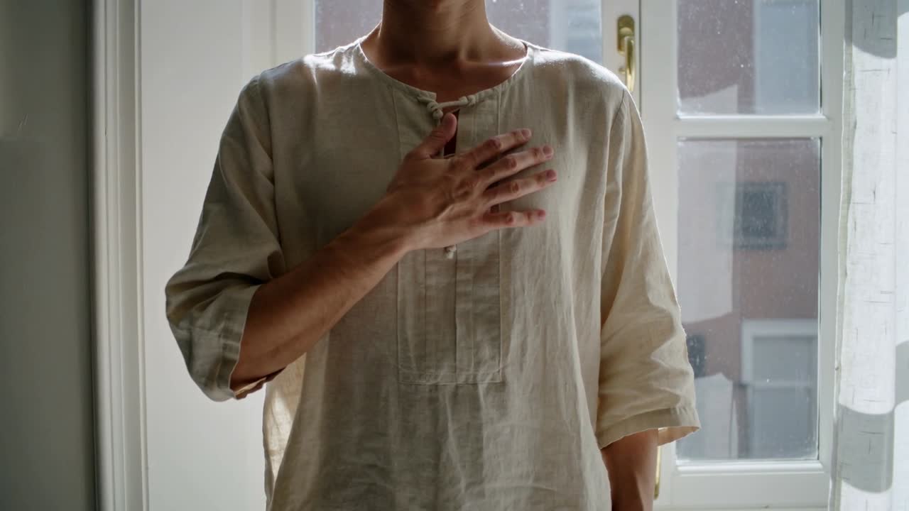 Donna che posiziona la mano destra sul petto davanti a una finestra alta, sistemando la tunica con la punta delle dita