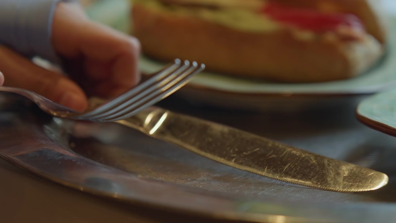 Primo piano di una mano che si allunga verso le posate su un tavolo con cibo sullo sfondo. Forchetta e coltello poggiano su piatti di metallo, riflettendo la luce delicatamente mentre rimangono immobili. La scena presenta un ambiente interno con luce soffusa.