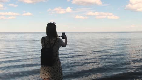 back view portrait of woman making selfie in the background of beautiful seascape using a smartphone and the monopod at sunset, slow motion