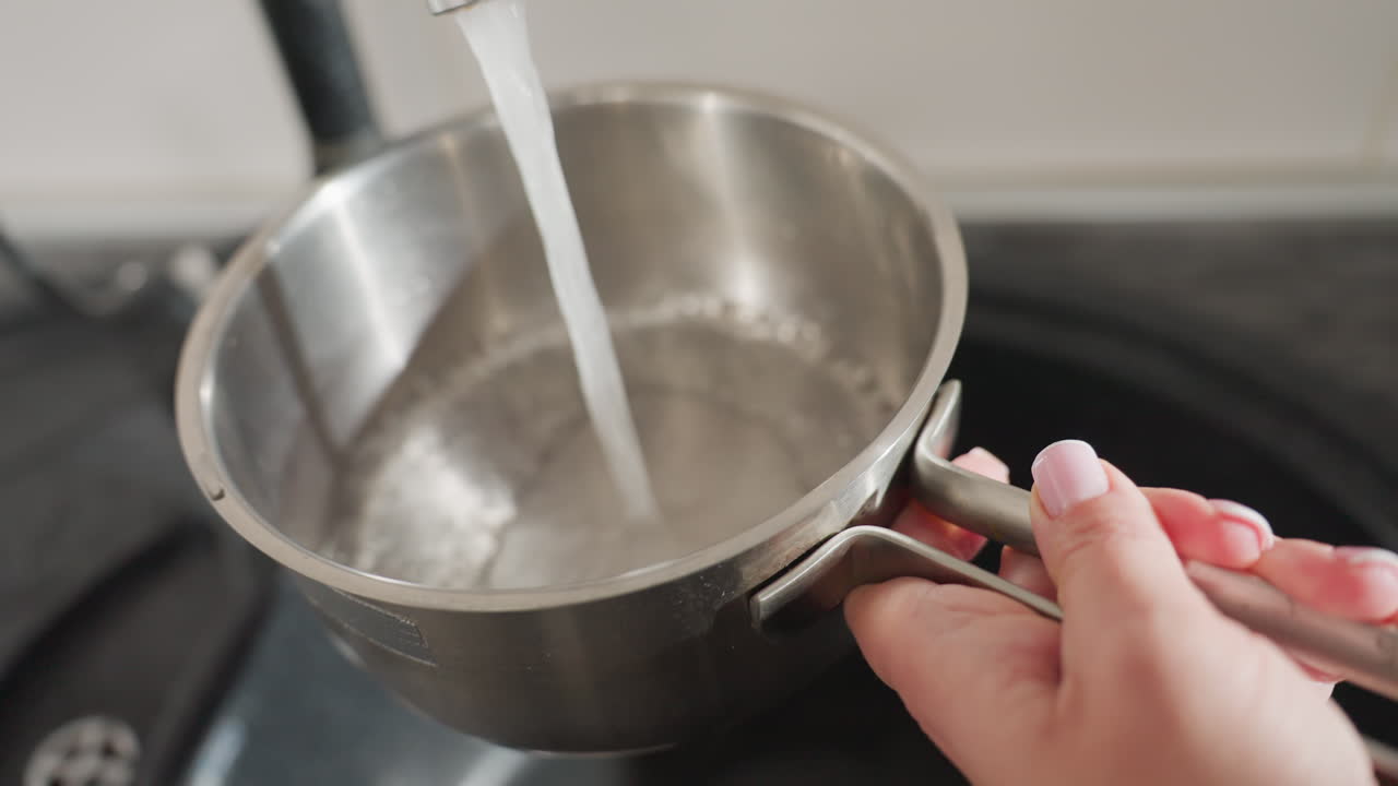 Close up of lady turning on kitchen tap to collect water into stainless steel pan, focusing on delicate hands with pink polished nails and casual jewelry, with modern clean kitchen setting