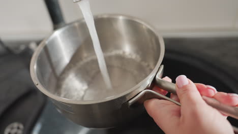 close up of lady turning on kitchen tap to collect water into stainless steel pan, focusing on delicate hands with pink polished nails and casual jewelry, with modern clean kitchen setting