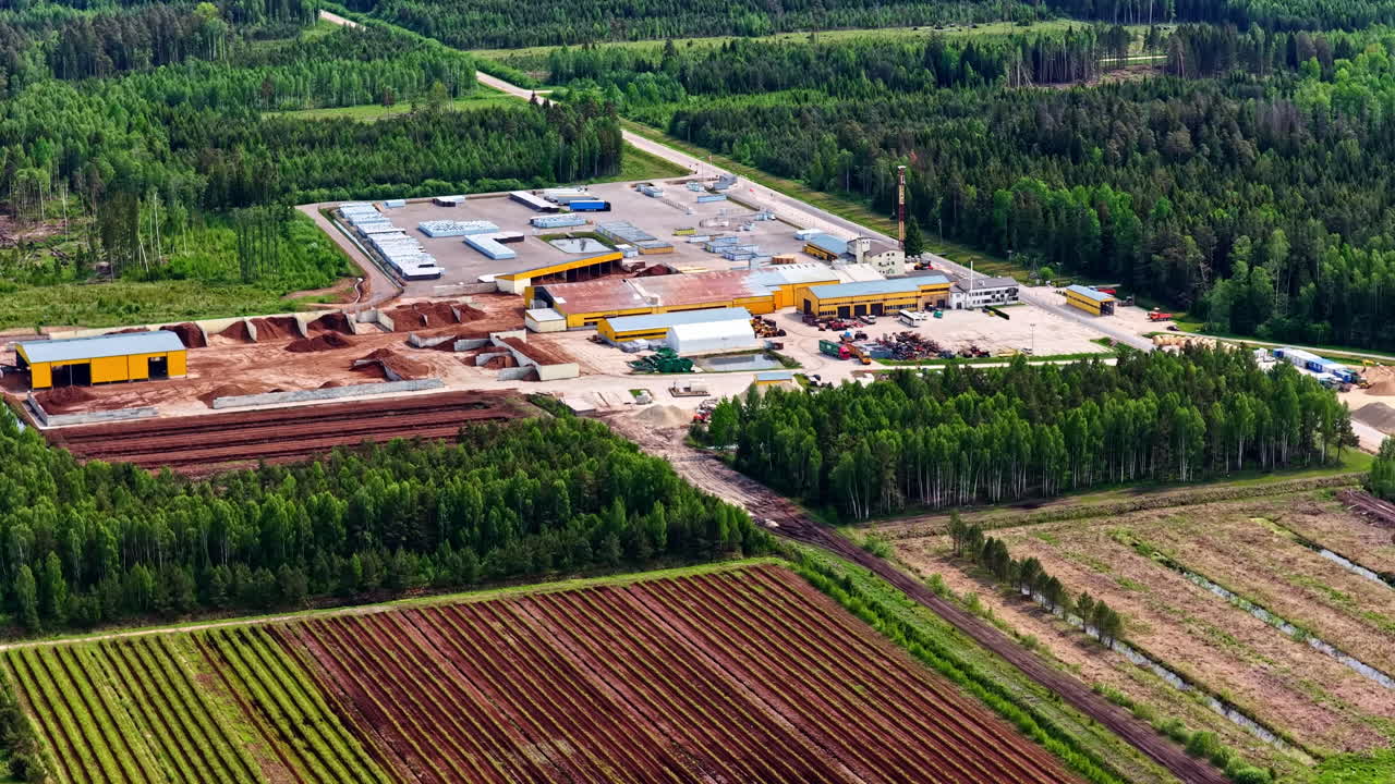 Drone view of an industrial facility surrounded by forest and farmland, showing warehouses, machinery zones, storage areas, and organized production infrastructure
