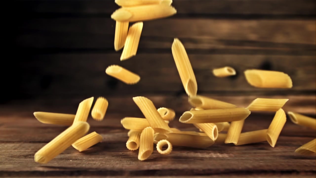 Pasta penne cruda che cade su un tavolo di legno rustico al rallentatore