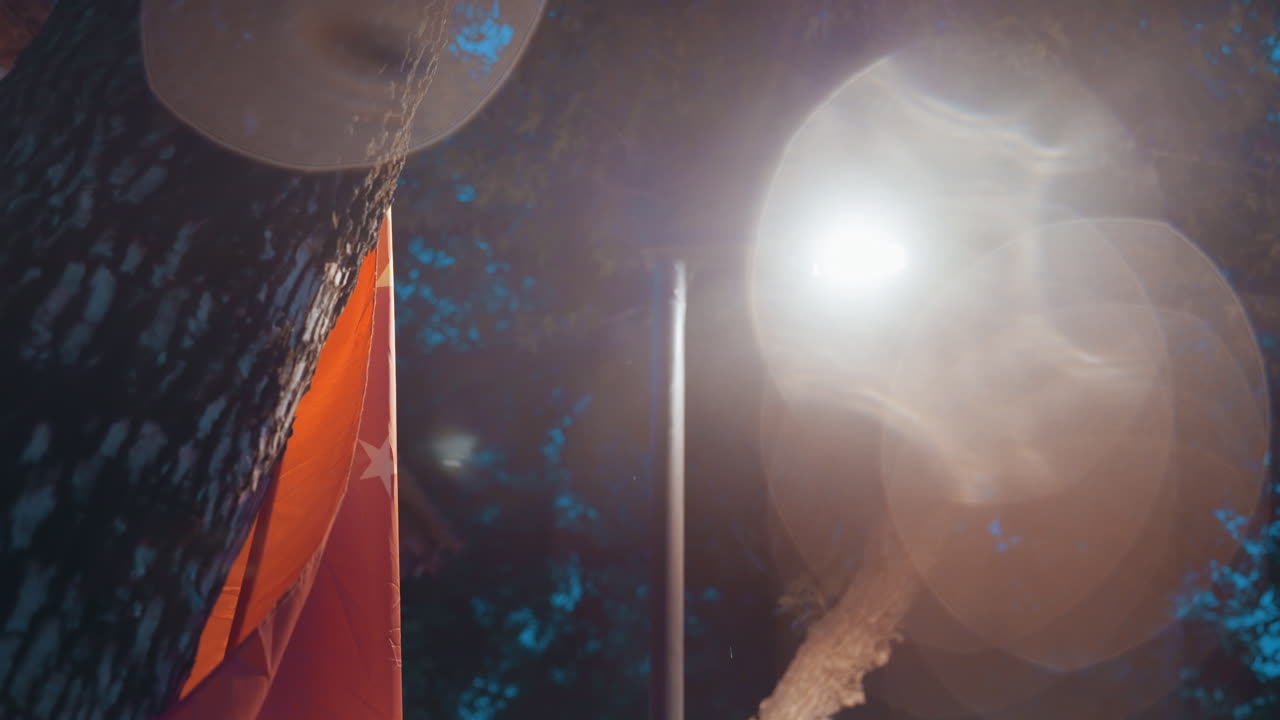 Primo piano di bandiera rossa e lampione notturno che sventolano sopra le grondaie, intenso flare dell'obiettivo e bokeh circolare, caldo bagliore e leggera foschia, trama del tessuto visibile contro un tranquillo profilo urbano di tetti, evoca nostalgia e solitudine.