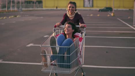 Fashion-Funny-Young-Hipster-Teen-Girls-Having-Fun-At-The-Shopping-Mall-Parking,-Riding-In-Shopping-Cart-Holding-The-American-Flag