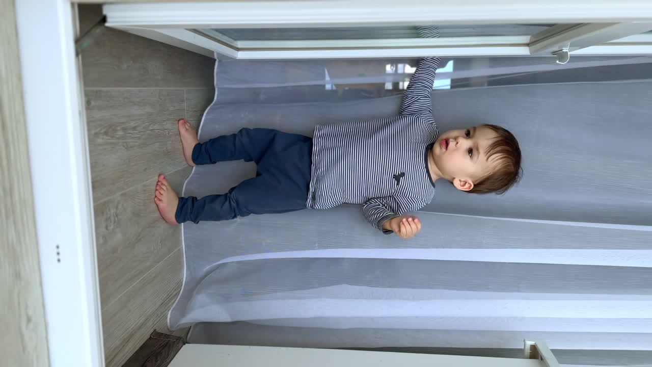 Sweet Caucasian baby boy stands at the balcony door and white curtain. Curious kid moves the door to and fro. Vertical screen.