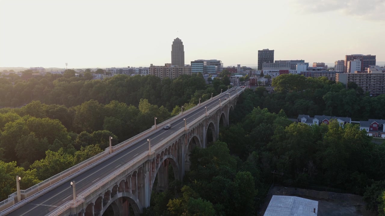 Il ponte Albertus Meyers in cemento ad arco trasporta auto sopra fitte cime di alberi verdi verso il centro di Allentown, Pennsylvania; la PPL Tower si erge sopra lo skyline della città al tramonto dorato. Ripresa aerea in discesa con drone