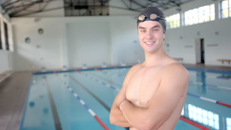 standing by indoor pool, male swimmer in swim cap and goggles smiling confidently, copy space