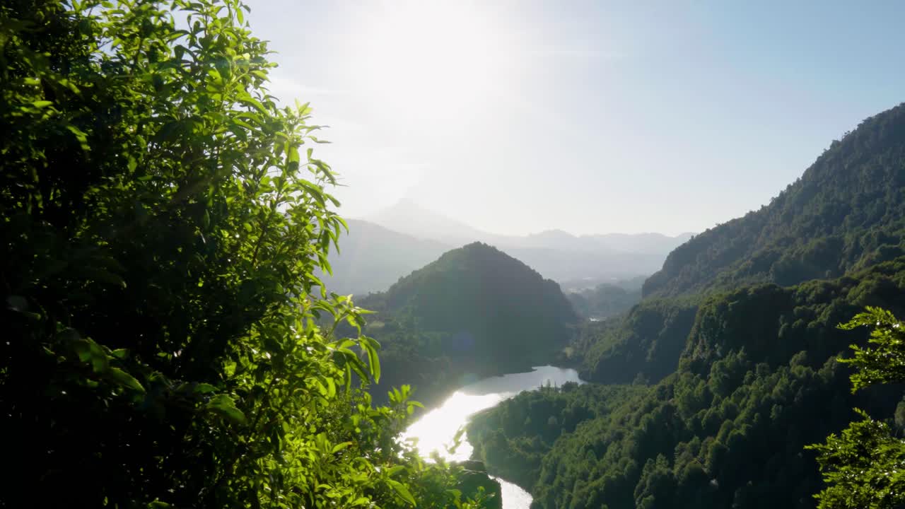 Sun beams between foreground bushes revealing river surrounded by mountains covered in green woods, El Leon viewpoint, Chile
