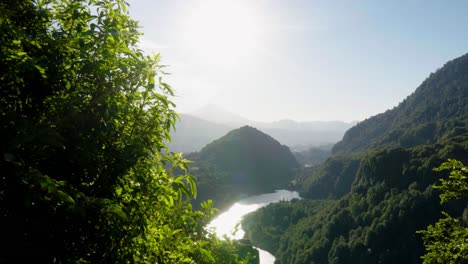sun beams between foreground bushes revealing river surrounded by mountains covered in green woods, el leon viewpoint, chile