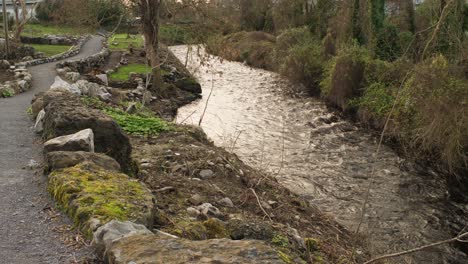 static shot of a flowing river in the city of gort, green vegetation, and a stone path on sunset