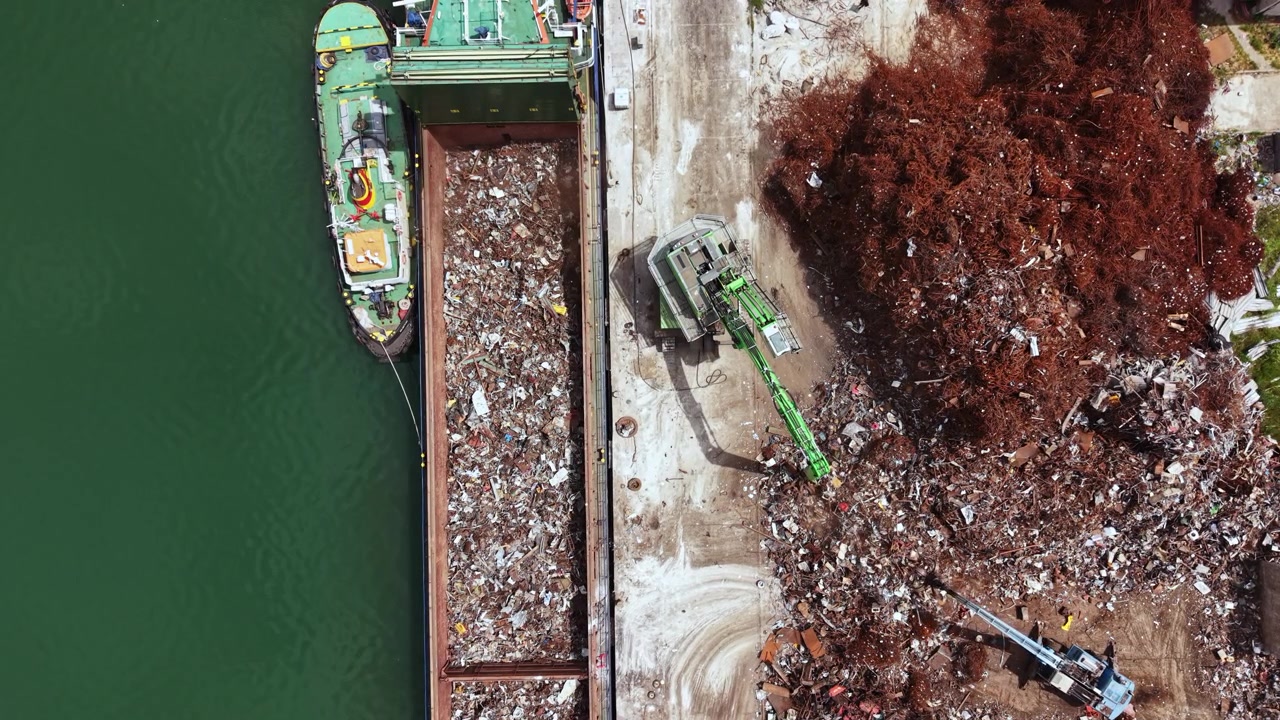 Grande nave che scarica rifiuti presso un impianto di riciclaggio vicino a un fiume in un'area urbana trafficata durante le ore diurne.