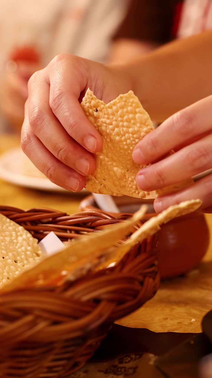Close-up of hands breaking crispy Indian papadum bread over a basket in warm restaurant lighting.