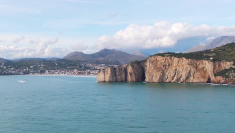 Water-view-of-Montagna-Spaccata-outcropping,-motorboat,-Gaeta-downtown-and-mountains-in-background-on-sunny-day,-Italy,-overhead-aerial