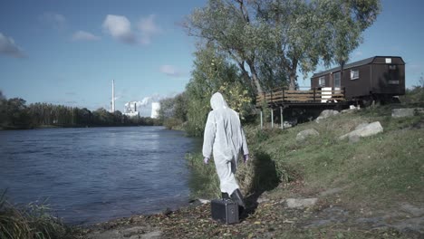 mujer con traje protector caminando hacia el río para hacer una muestra de agua, sosteniendo un tubo de ensayo y un estuche, siguiendo el tiro de la muñeca