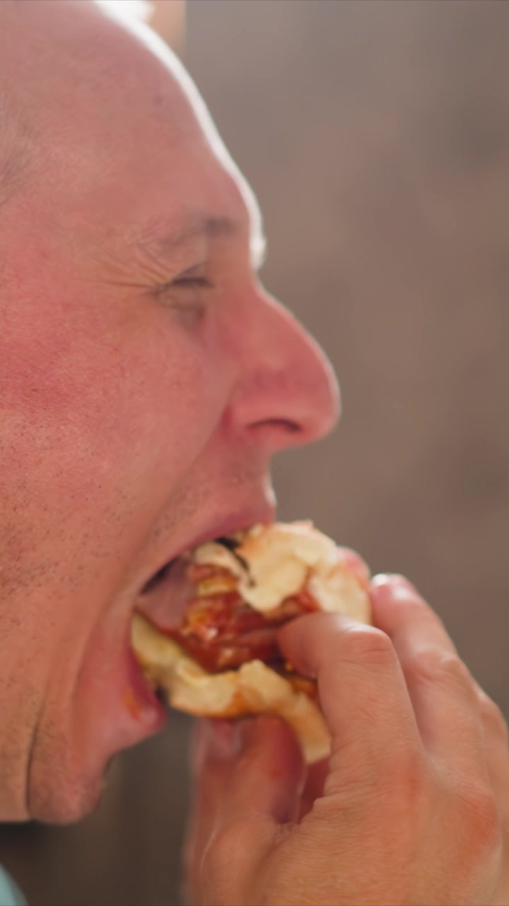 Primo piano di un uomo che mangia cibo con la mano visibile. Il soggetto tiene e addenta vari cibi, inclusi panini o hamburger, durante la sequenza. L'illuminazione soffusa crea un aspetto naturale della pelle.