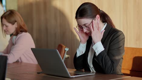 Close-up-of-a-tired-brunette-girl-in-business-clothes-loses-her-temples-and-adjusts-her-glasses-while-working-in-front-of-a-laptop-in-a-modern-sunny-office