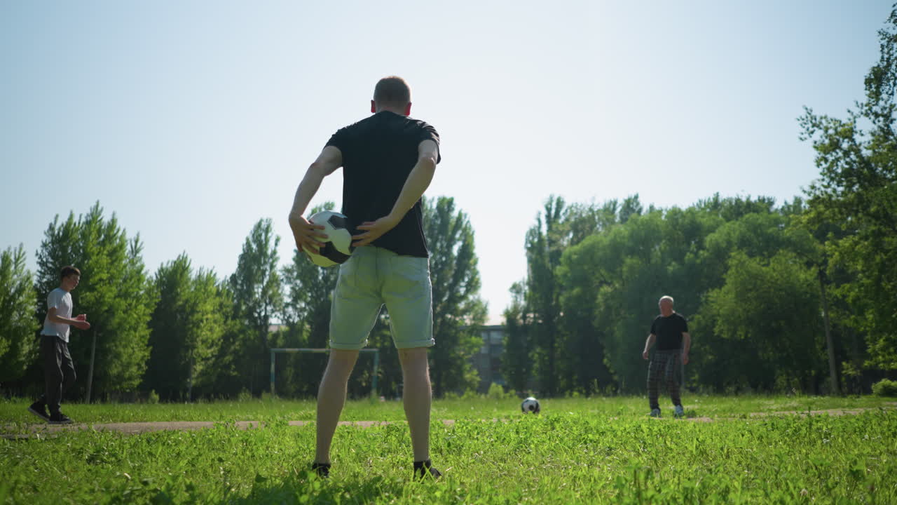 A close-up of a man rotating a soccer ball in his hands, with a view of people engaging in a soccer game in the background