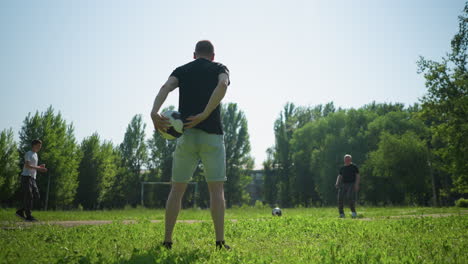 a close-up of a man rotating a soccer ball in his hands, with a view of people engaging in a soccer game in the background