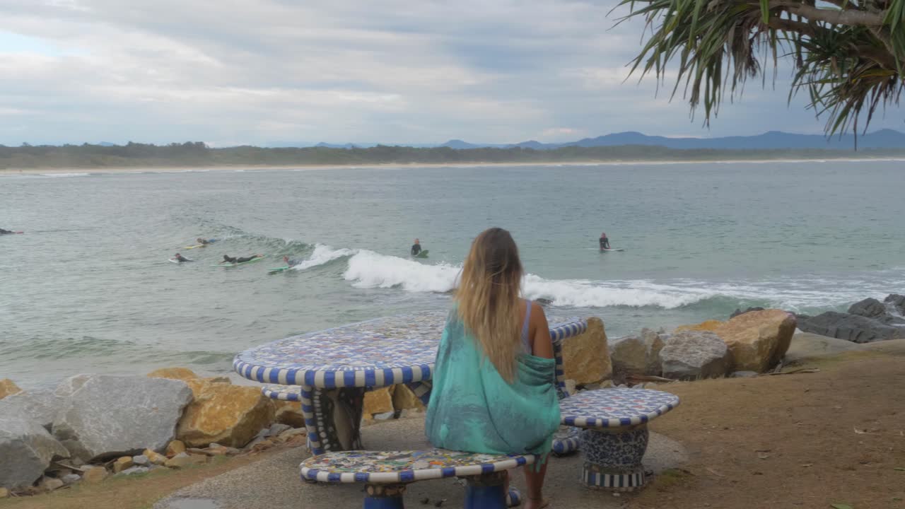 vista trasera de una mujer mirando a los surfistas en el océano mientras se sienta en una mesa de picnic - parque en el mirador de scotts head, nsw, australia