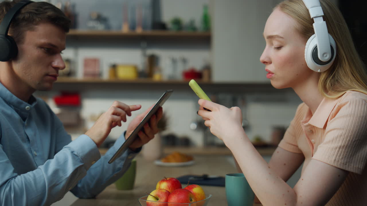 una pareja usando aparatos por separado disfrutando de música en auriculares en un primer plano de la cocina.