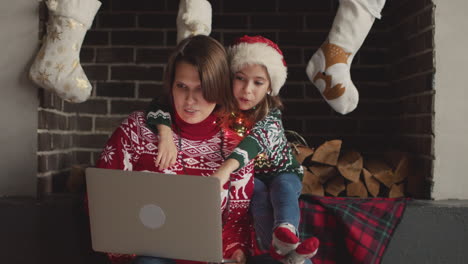 feliz madre e hija en suéteres navideños y con luces alrededor del cuello sentadas en el sofá mientras usan una computadora portátil