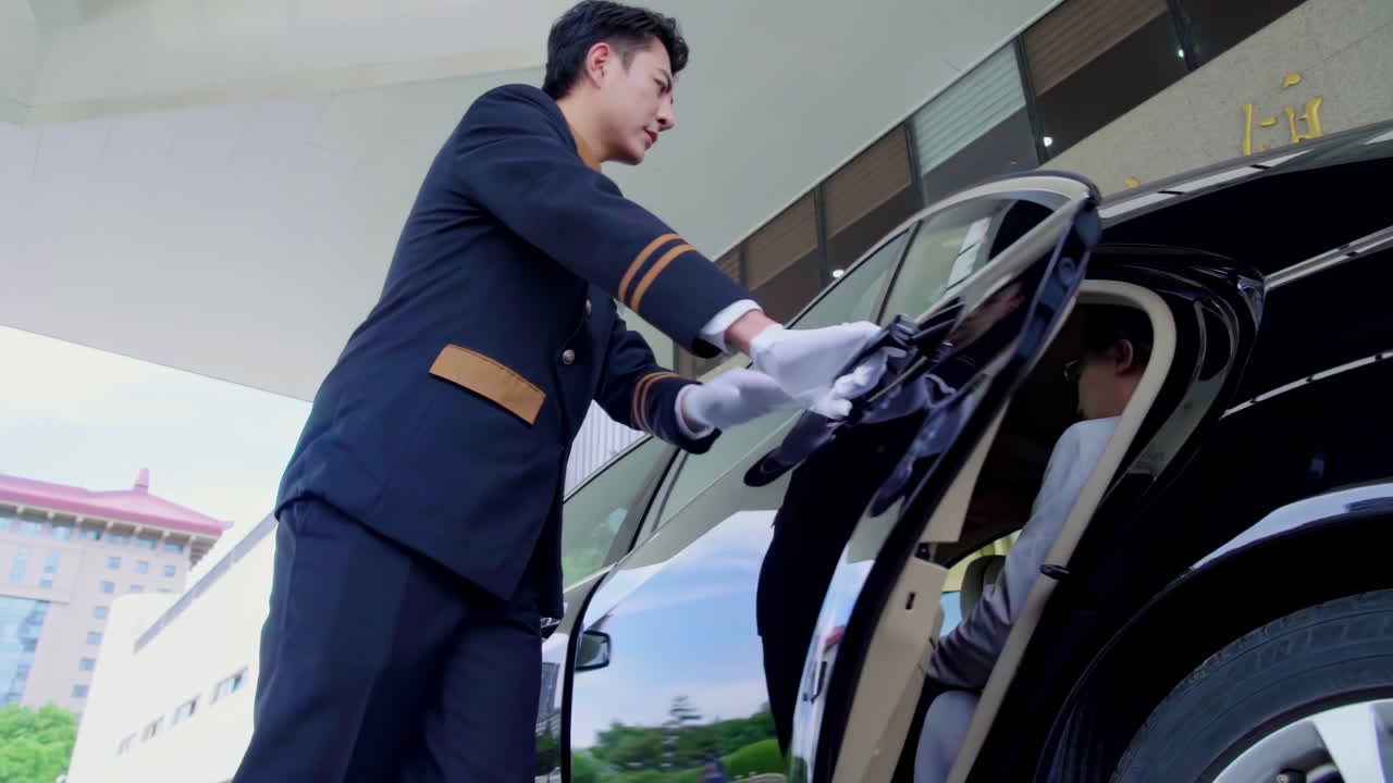Smiling porter in uniform opening door of taxi cab and greeting female guest arriving at hotel. Service-minded doorman opening car door for guest