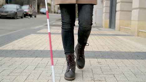 vista media de una mujer ciega con abrigo marrón y botas negras caminando con un bastón en la calle