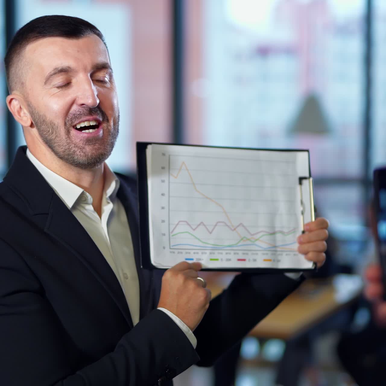 Hand with a phone taking video of a man with chart. Handsome man showing graph patterns and explaining them to the camera