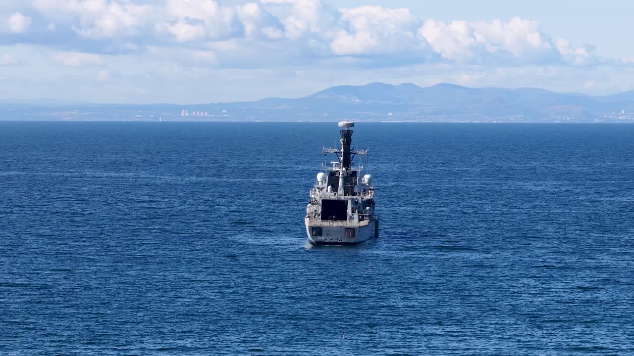 Military vessel advances steadily across blue ocean under bright daylight, distant hills visible, static camera
