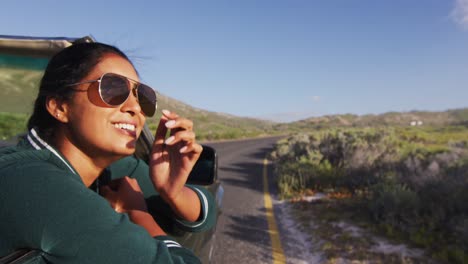 Mixed-race-woman-driving-on-sunny-day-in-convertible-car-leaning-on-door-smiling