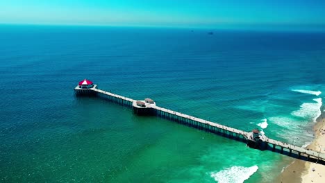 alto drone vista volando alrededor del lado del muelle en huntington beach california con una gran vista de las montañas, playa, olas, surf e incluso algunas personas caminando alrededor
