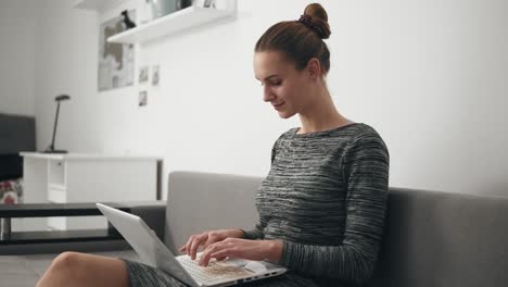 Young-woman-at-home-sitting-on-the-sofa,-she-is-working-with-a-laptop-and-typing-text-looking-at-the-screen