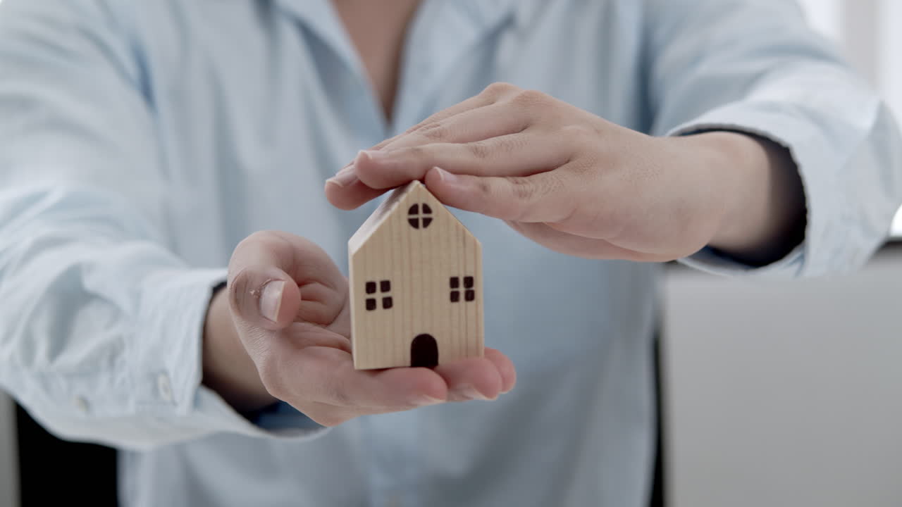 una persona coloca cuidadosamente sus manos sobre un modelo de casa de madera que simboliza la protección del hogar.