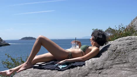 Girl-sunbathing-on-rock-at-beach-and-drinking-beverage