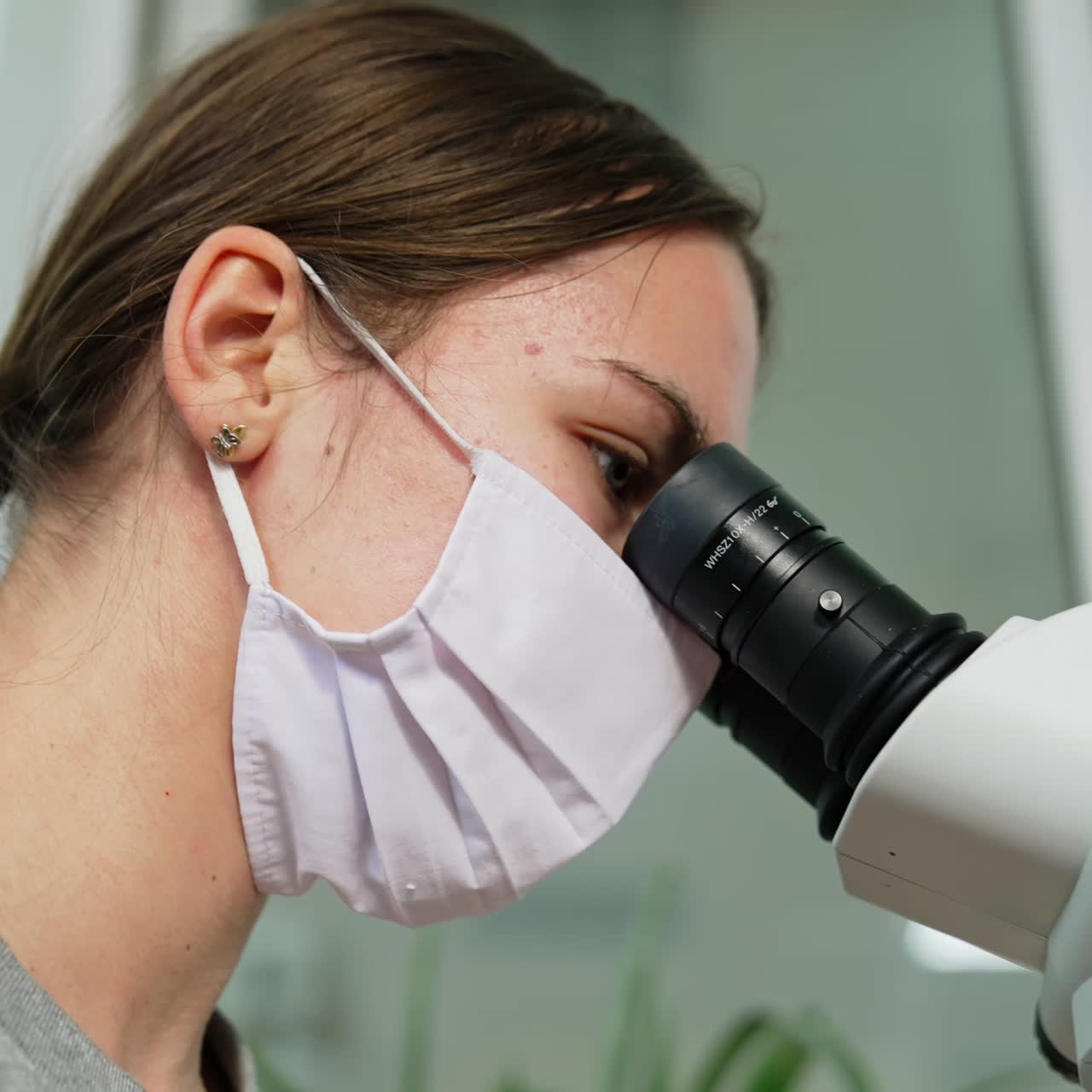 Brunette girl in mask looks carefully at microscope binoculars. Ophthalmic student practicing. Low angle view