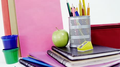 colored pencils in pen holder with pile of books, apple and stationery