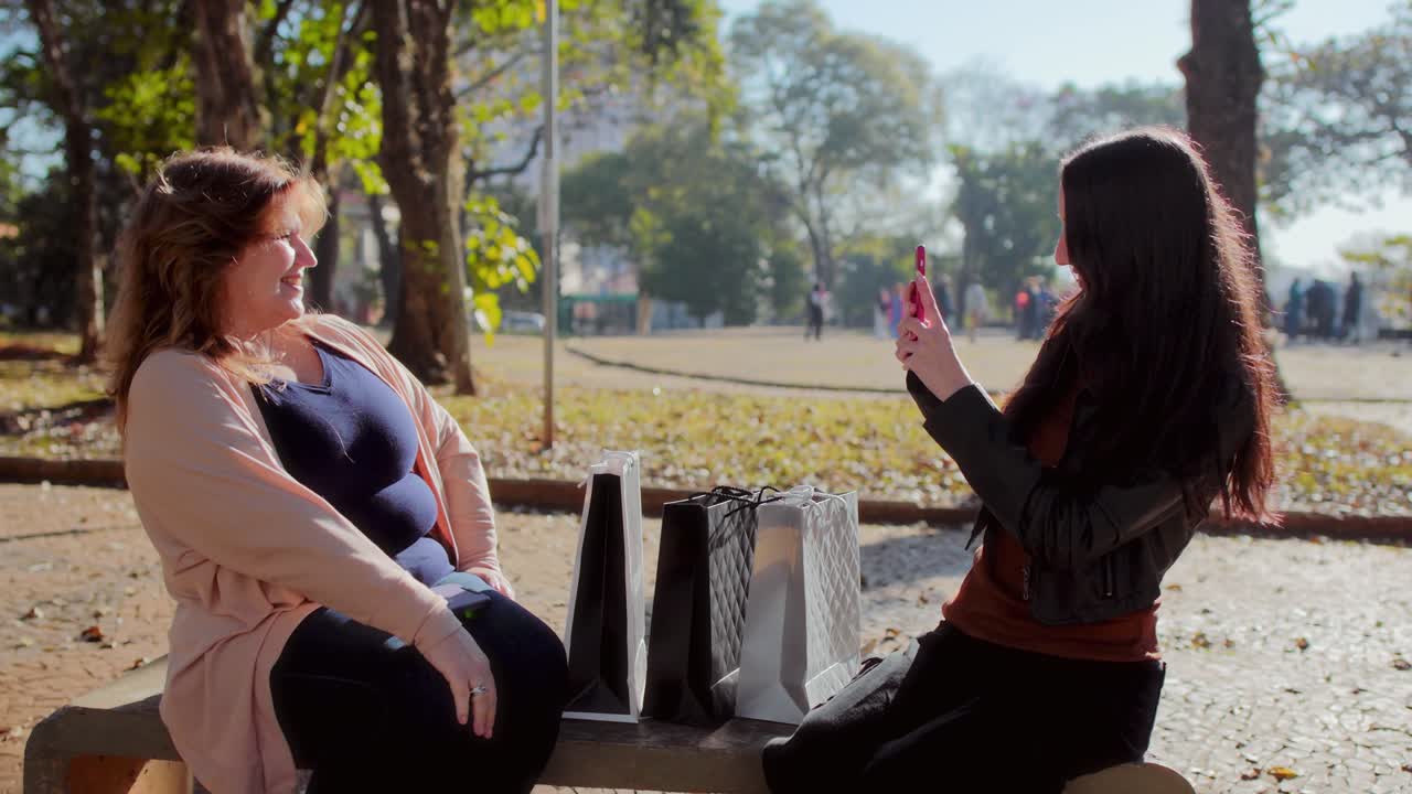 Two women sitting on a park bench with shopping bags, talking and interacting with a smartphone