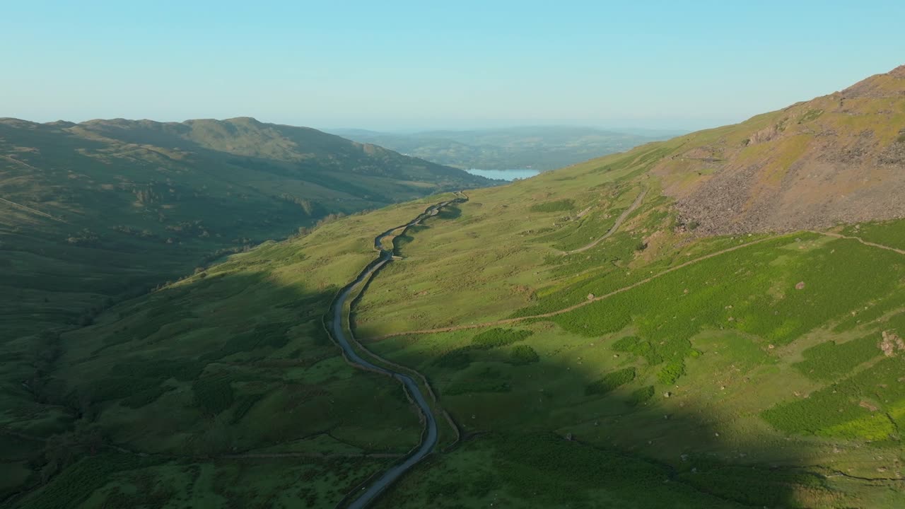 Mountain pass road snaking out of valley shadow into sunrise light towards distant lake Windermere. Summer. Kirkstone Pass, Lake District, Cumbria, UK