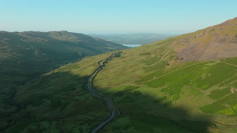 mountain pass road snaking out of valley shadow into sunrise light towards distant lake windermere. summer. kirkstone pass, lake district, cumbria, uk