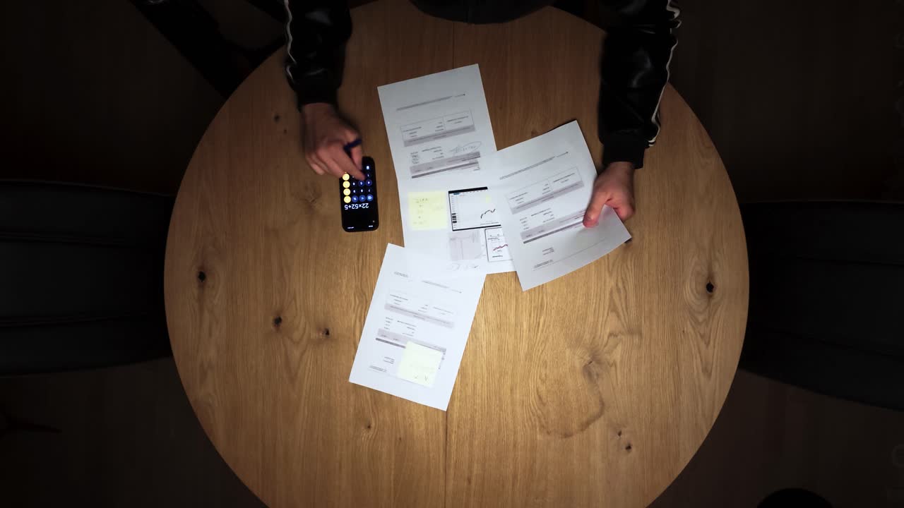 A person reviews invoices and calculates expenses on a wooden table using a calculator, representing financial management and budgeting
