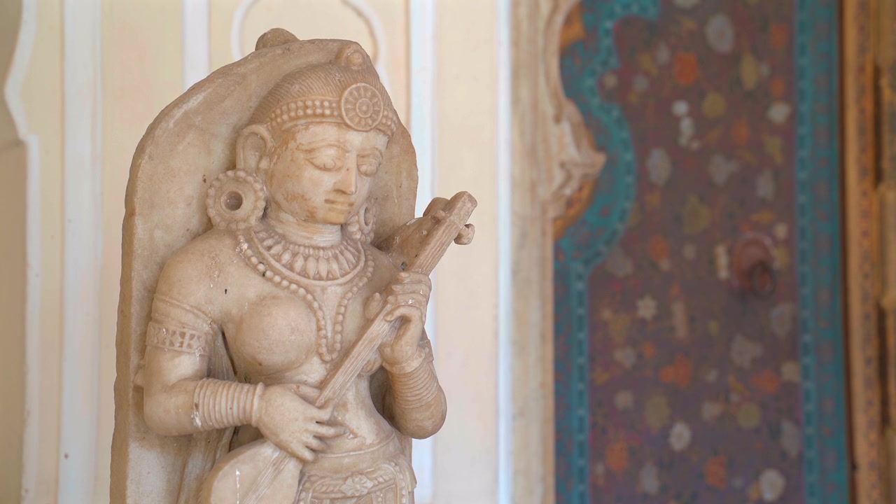 Inquadratura statica in primo piano medio di un'antica scultura in pietra raffigurante una figura femminile che suona uno strumento musicale davanti a un arco decorato a Jaipur, Rajasthan, India