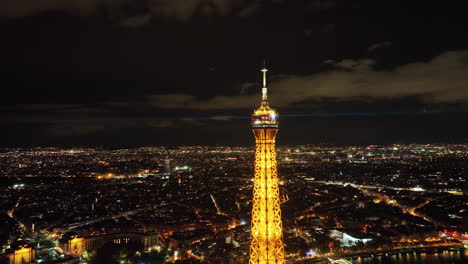 Cumbre-De-La-Torre-Eiffel-Con-Haces-De-Luz-Automatizados-Que-Brillan-Por-La-Noche,-Paisaje-Urbano-De-París-Y-Horizontes-En-El-Fondo,-Vista-Aérea